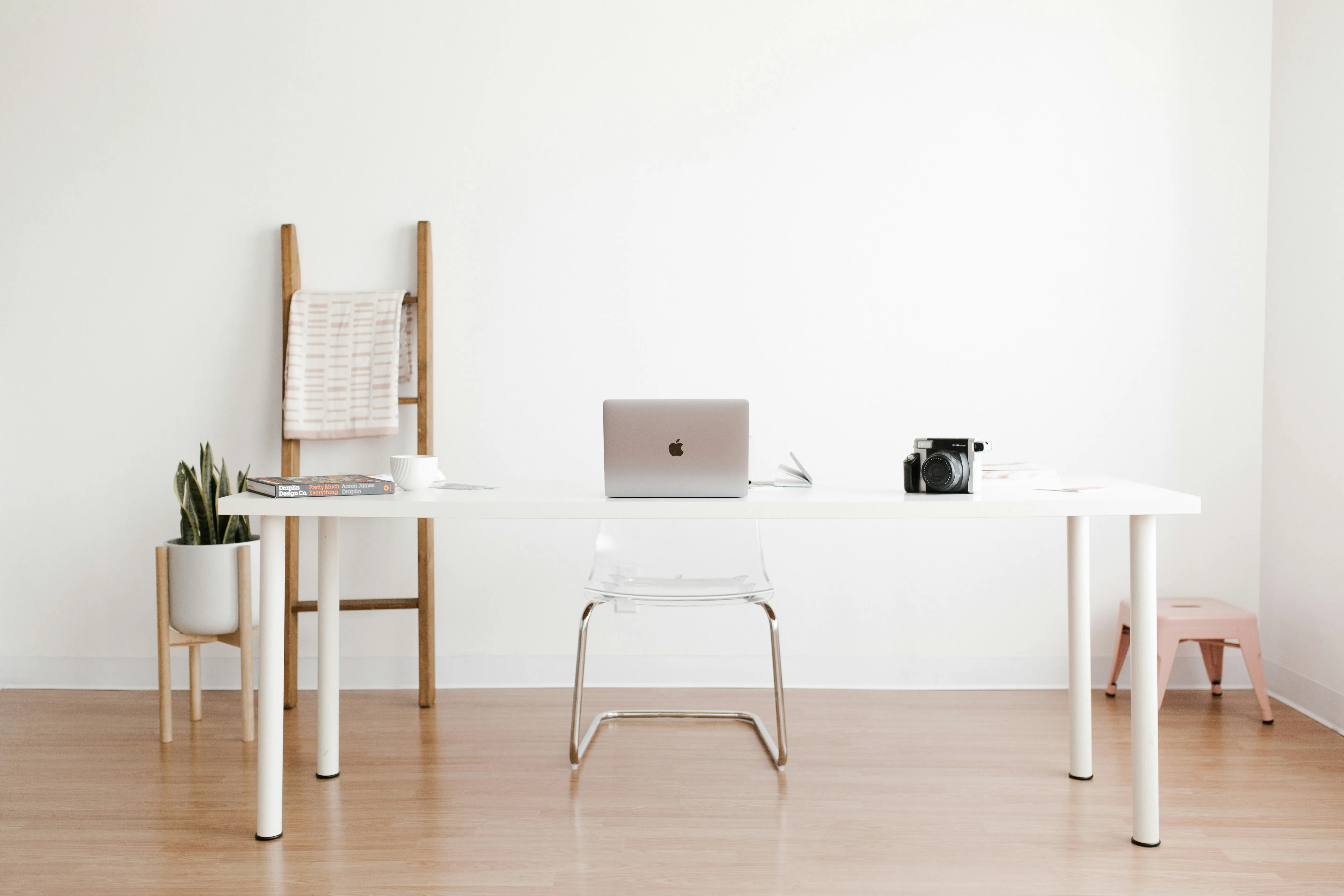 Minimalist home office setup with a white desk, MacBook, vintage camera, clear acrylic chair, potted snake plant, and wooden ladder against a white wall